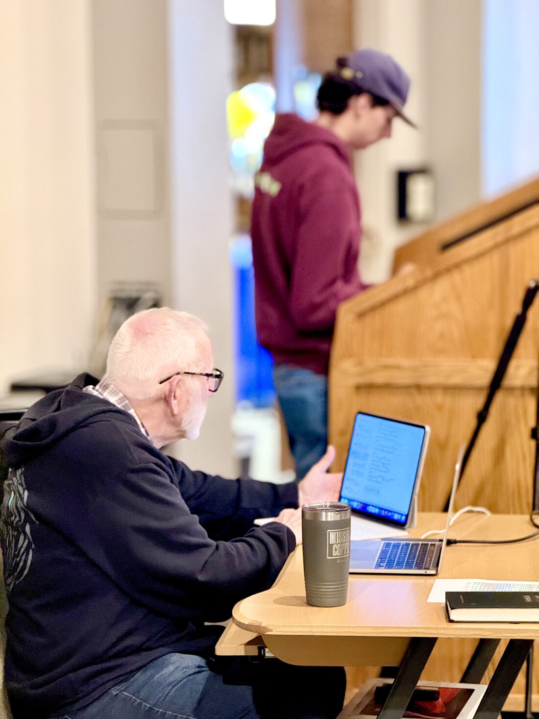 A Cornerstone volunteer checking a tablet and laptop at the production table while another sets up the sound booth